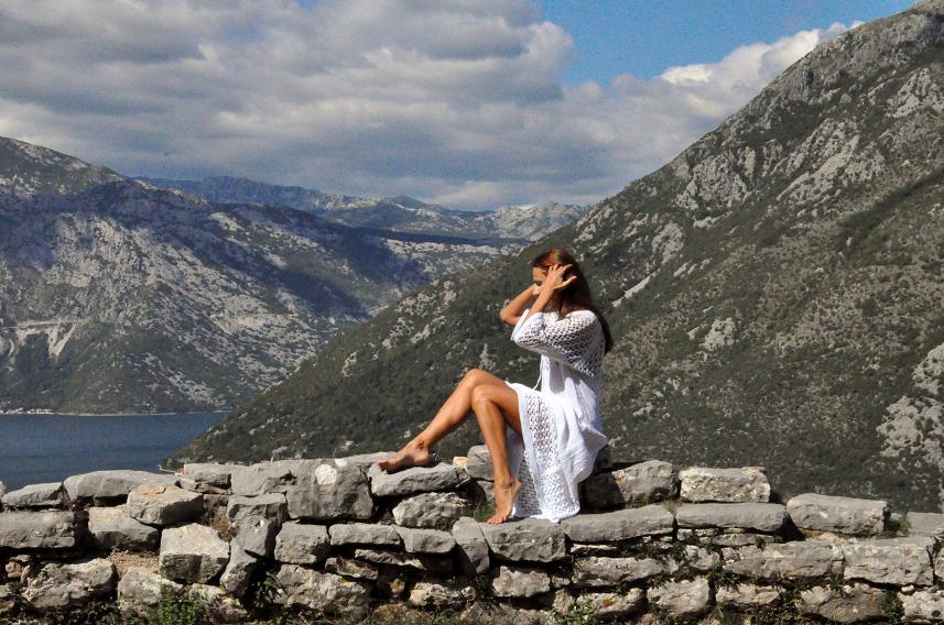 Woman in white sitting on stone wall above Kotor Bay